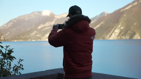 Male-Wearing-Red-Hooded-Winter-Jacket-Taking-Photo-Of-Lake-With-Smartphone-From-Viewpoint