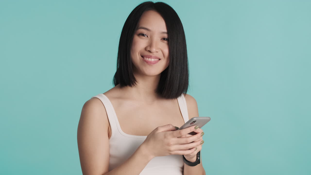 Asian woman texting on smartphone and smiling at the camera.
