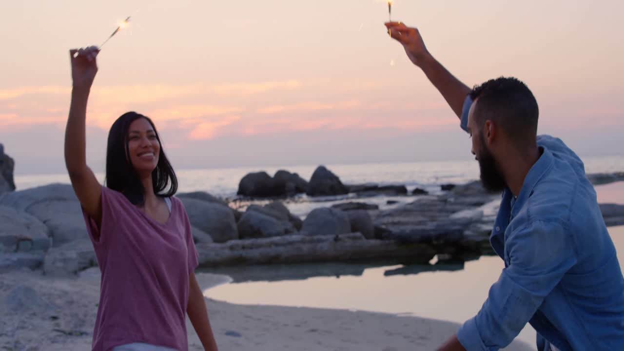 feliz pareja joven de raza mixta divirtiéndose con chispas en la playa 4k