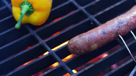 hand picks up yellow pepper from grill with sausages cooking over fire at campsite