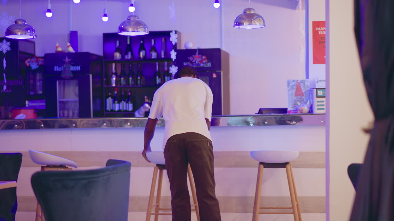 Man cleaning bar counter in modern lounge with purple lighting, alcohol bottles on shelves, and stylish chairs in the background. A professional setting for bar and hospitality services