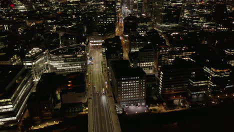 Backwards-reveal-of-vehicles-driving-on-wide-multilane-street-between-multistorey-buildings.-Aerial-view-of-city-at-night.-London,-UK