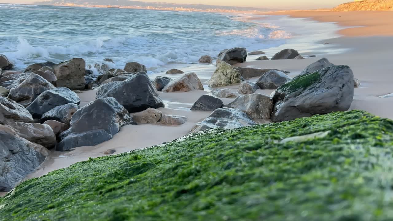 Atlanterhavsbølger ruller ind på en sandet og stenet strand med solbelyst baggrund.