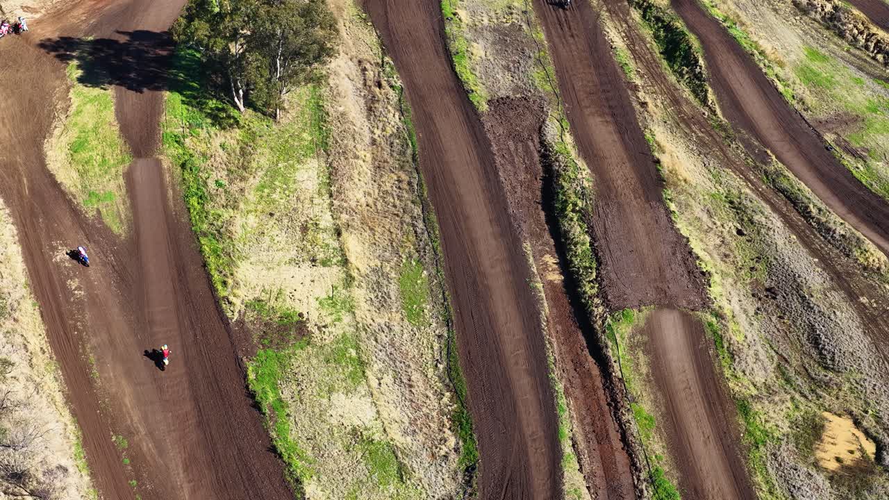 Multiple motorbikes speed along winding dirt track, captured in bright daylight from overhead drone perspective