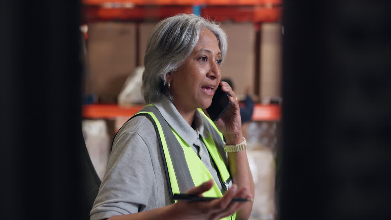 Mature Woman in Safety Vest Talking on Phone in Warehouse