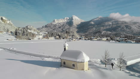 aerial drone shot showing a snowy landscape in in the sunshine of the tyrol, austria and a snow covered tiny chapel in lechaschau
