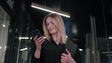 mujer de negocios sonriente mirando el teléfono celular en el interior. mujer de negocios sorprendida leyendo un mensaje en el teléfono móvil en el pasillo de la oficina. mujer mirando la pantalla del teléfono inteligente en el centro de negocios. oficina caminando