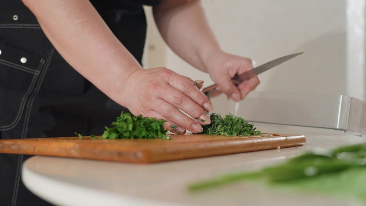 Mani che tagliano, raccolgono e trasferiscono erbe fresche verdi su un tagliere di legno mentre il coltello affetta verso il basso e le dita uniscono il mucchio per cucinare e guarnire