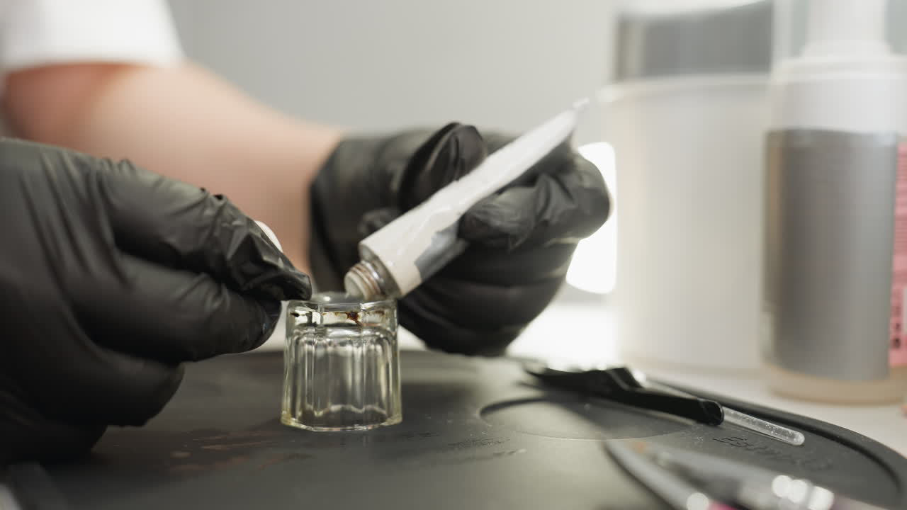 Beautician wearing black gloves dispenses product from tube into small glass jar on black silicone mat surrounded by cosmetic tools, preparing substance for professional beauty treatment session
