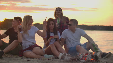 los hombres y mujeres jóvenes están sentados alrededor de una fogata en la costa del río. están bailando con cerveza y hablando entre sí al atardecer en la noche de verano.