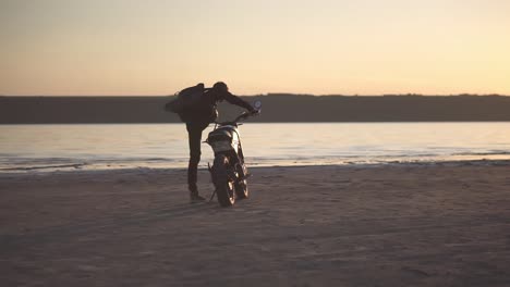 rare view of a biker walking to his bike, sits on it. the black bike parked close to the water with sunset view