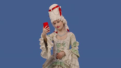 retrato de una dama de la corte con un vestido de encaje vintage blanco y una peluca tomando una selfie en un teléfono móvil. una mujer joven posando en un estudio con fondo de pantalla azul. de cerca. cámara lenta lista 59.94fps