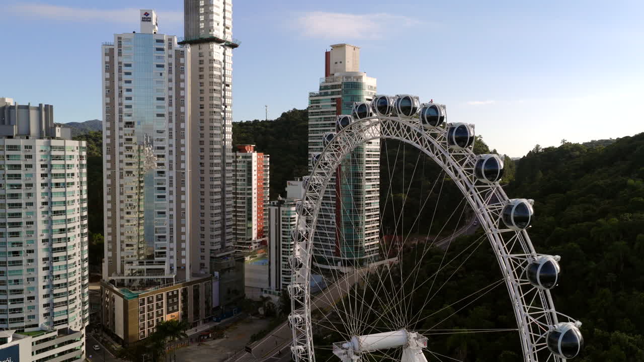 Towering over Barra Norte skyline, prominent ferris wheel rising against bright sky, highlighting urban landscape of Balneario Camboriu, Santa Catarina, Brazil, drone pushing in