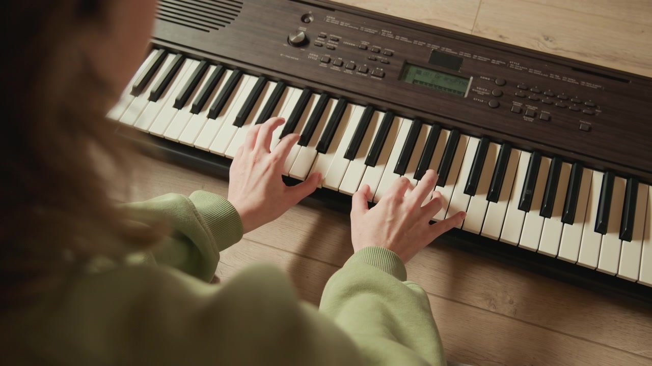 Højvinkelindramning viser en siddende person, der spiller elektronisk keyboard på trægulv, begge hænder trykker på tangenterne med buede fingre, afslappede håndled og jævn bevægelse under fokuseret øvelse.