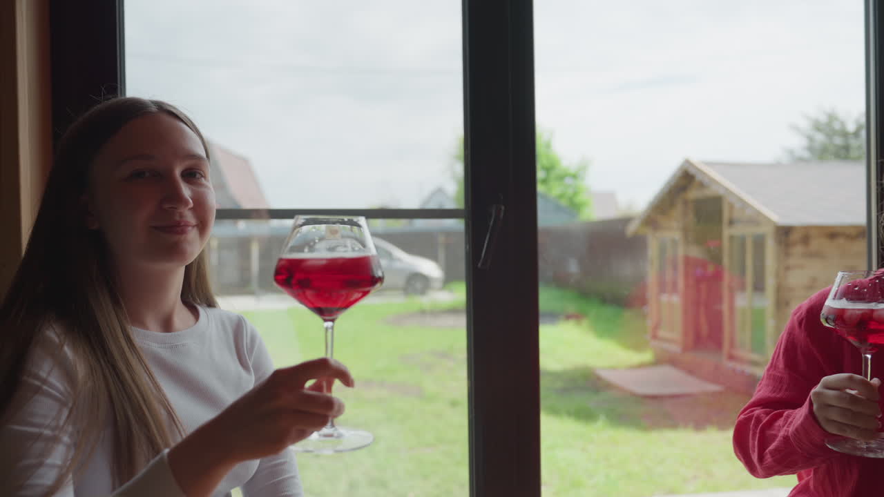 Family enjoying joyful moment as they raise glasses filled with red drink and ice cubes near large window, surrounded by daylight and backyard view, sharing cheerful toast together indoors