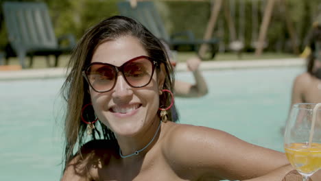 close up of attractive woman in swimming pool posing for camera, smiling and drinking beverage while her friends are having fun behind