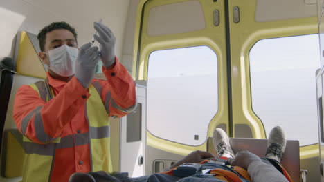 male paramedic with face mask preparing a syringe for an injection while an injured american patient lying on the ambulance strecher