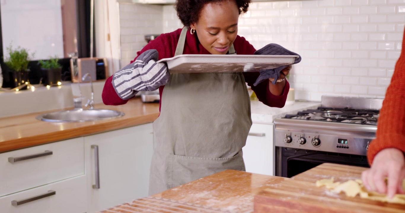 Female friends baking, oven timer dinging, woman in maroon with mitts presenting cookies to partner