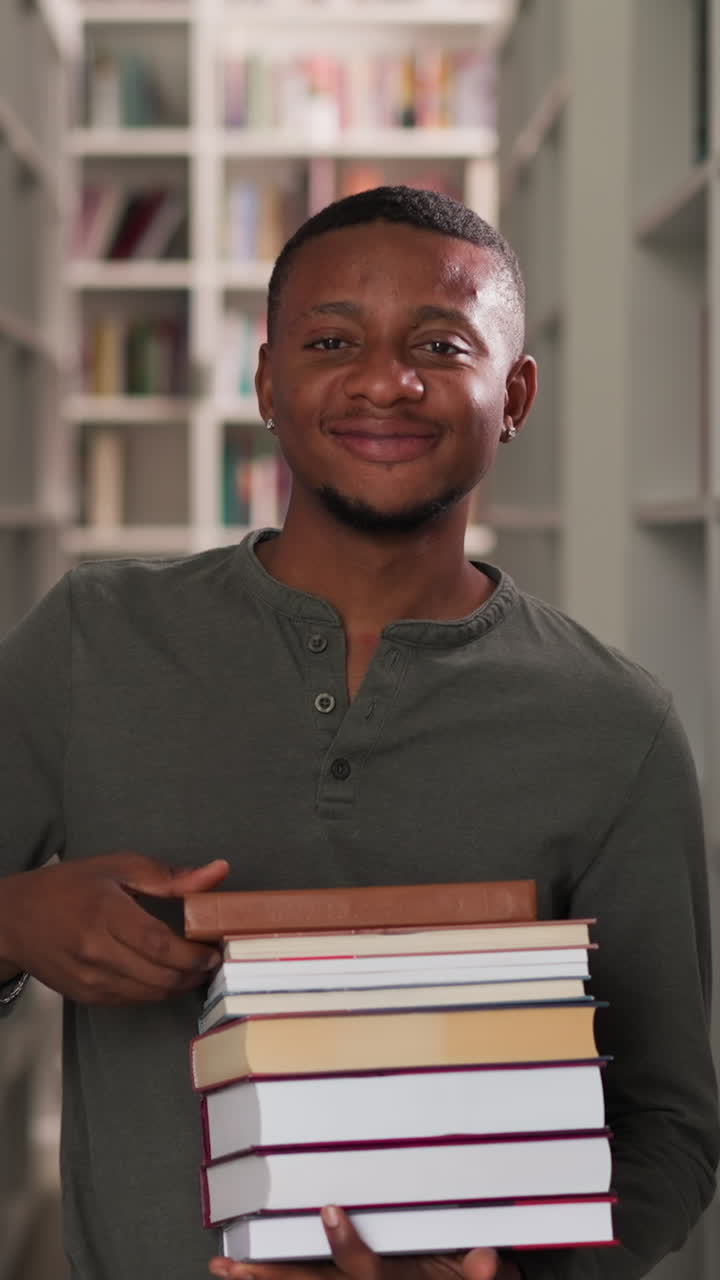 un hombre feliz sostiene una pila de libros en una biblioteca pública. un bibliotecario afroamericano asente con la cabeza llevando una pila de literatura entre las estanterías. un estudiante muestra folio