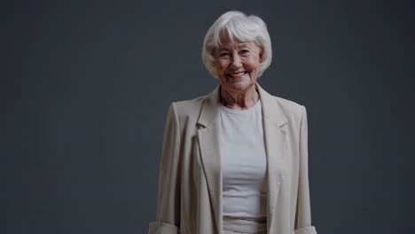 confident senior businesswoman with white hair wearing light casual clothing radiating optimism, smiling warmly while looking directly at camera against neutral gray studio background