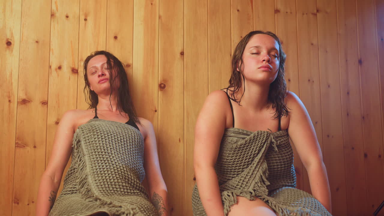 two women wrapped in absorbent towels sit against wooden sauna panels, cooling off after heat exposure, damp hair clinging to shoulders, beads of sweat glistening on skin