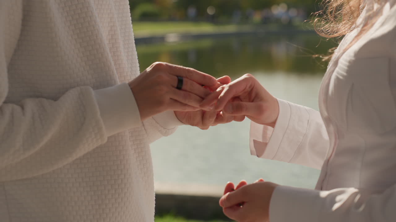 Closeup ring exchange between caucasian partners. Sunlit hands glide a band onto finger near a peaceful waterfront, detailed jewelry shot capturing tenderness, subtle emotion and symbolic promise