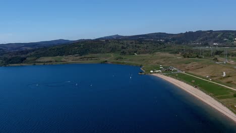 Lake-beach-with-people-sunbathing-and-bathing,-small-sailboats-sailing,-garden-and-walking-area-with-the-houses-in-the-background