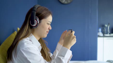 행복한 십대 여성은 헤드을 착용하고 침대에 스마트폰으로 온라인 게임을 하고 있으며, 여성은 스마트폰에서 게임을 잃었습니다.