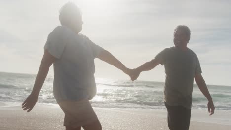 Feliz-Pareja-De-Ancianos-Hispanos-Tomados-De-La-Mano,-Caminando-Por-La-Playa-Al-Atardecer