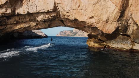 Toma-Aérea-Siguiendo-A-Un-Surfista-Eléctrico-Bajo-Un-Arco-De-Roca-Frente-A-La-Costa-De-España