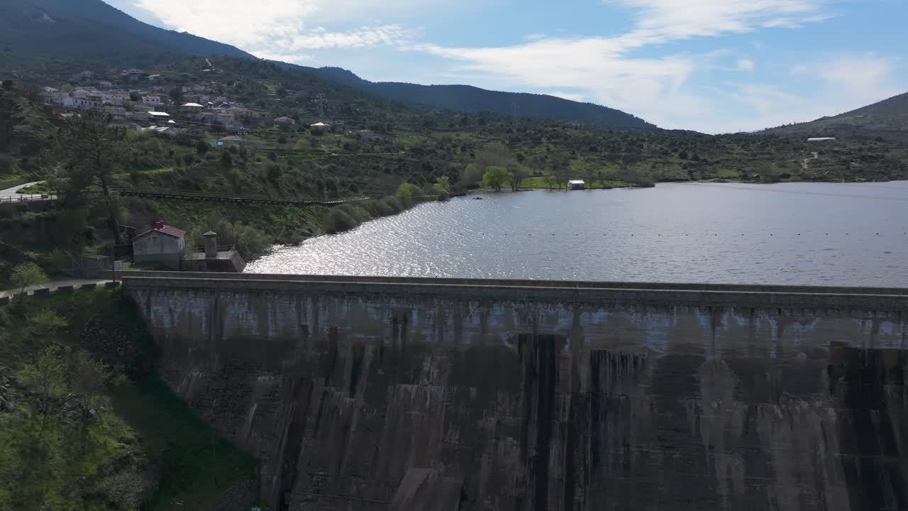 Lateral drone flight along a fully loaded dam wall. Sunlight reflects on the water's surface as we move, ending over a lush green area with two visible crossing paths.