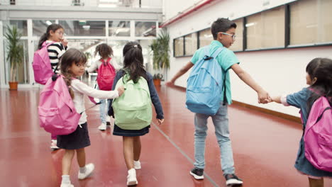 back view of happy multiethnic group of schoolkids with colorful backpacks walking along school corridor