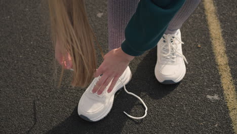 close-up of lady with flowing hair tying her white canvas sneakers on tarred road with yellow road markings, highlighting focus on shoelaces and athletic wear