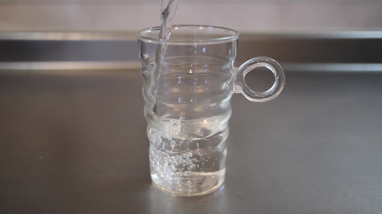toma completa de agua vertida en una taza de vidrio sobre una encimera de cocina metálica, concepto de salud y agua limpia, imágenes en cámara lenta