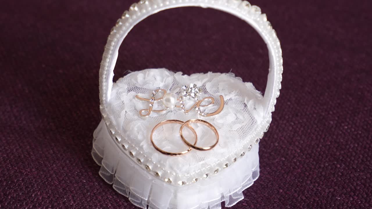 close up of two gold wedding rings resting on white lace heart basket with pearl and love charm