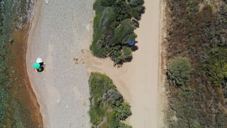 Vista-De-Pájaro-De-La-Playa-Idílica-Con-Turistas-Bajo-El-Paraguas-En-La-Orilla-Del-Mar-Durante-Un-Día-Cálido-Y-Soleado-En-Cerdeña,-Italia---Toma-Aérea-De-Drones