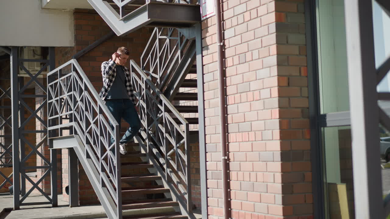 student descending outdoor metal staircase halts placing cigarette between lips lifts lighter sparks ember then inhales slowly while sunlight casts geometric shadows on brick facade and metal railing