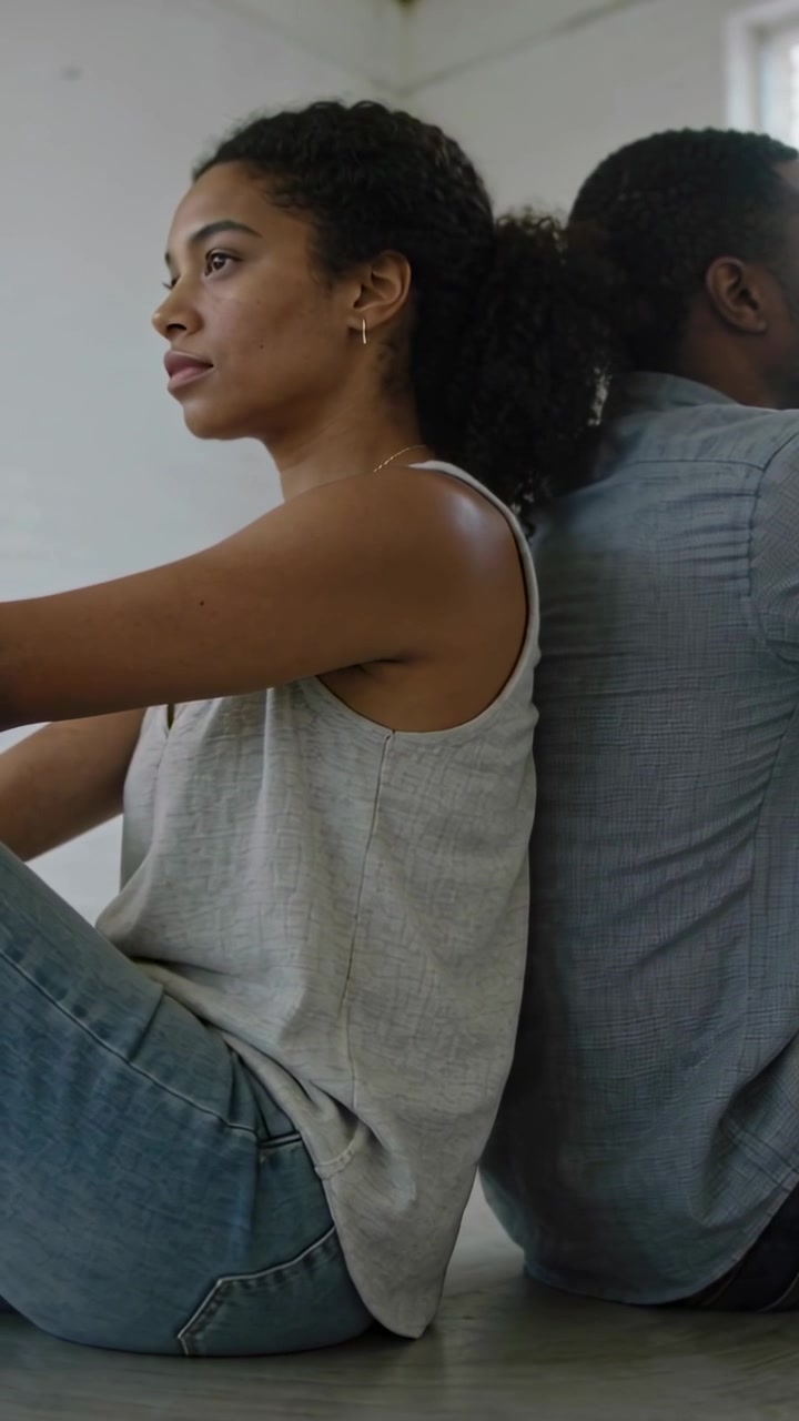 Vertical video: Sitting woman in gray top, jeans in sparse room shifting gaze to warm window light