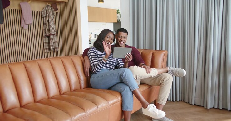 Pareja afroamericana sentada en un sofá color canela con ropa informal, usando una tableta durante una llamada y saludando