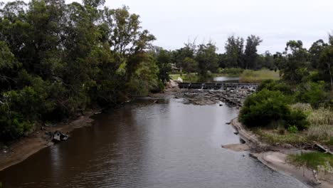 Aerial-video-touring-the-stream-with-a-dam-while-water-falls,-with-the-landscape-and-rocks-in-the-background