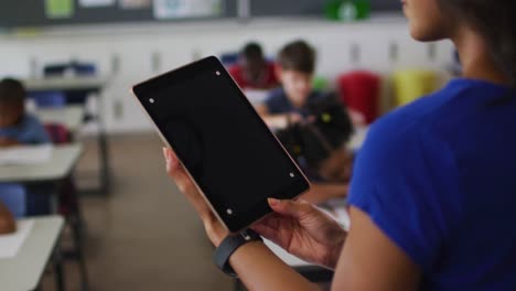 Midsection-of-mixed-race-teacher-using-tablet-with-copy-space-on-screen,-children-in-background