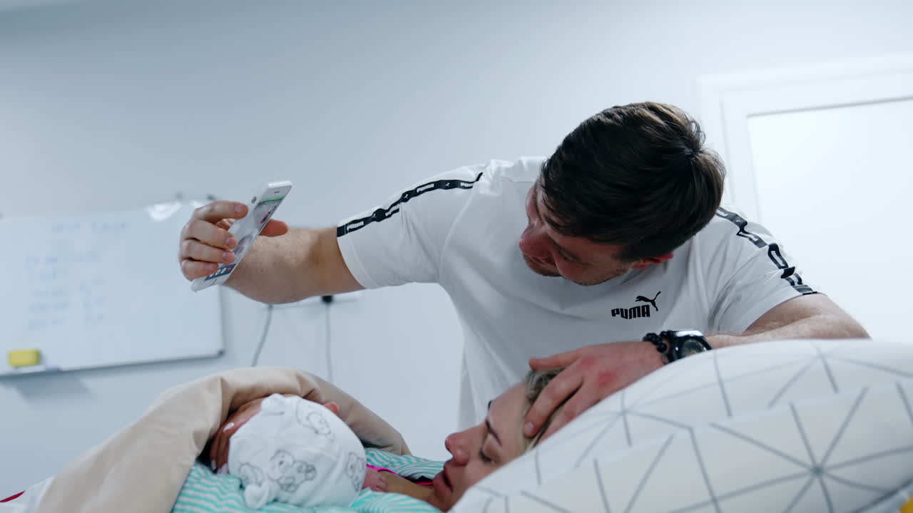 Happy man is holding a phone over his wife lying in bed. Woman talks to someone through video chat showing her newborn baby.