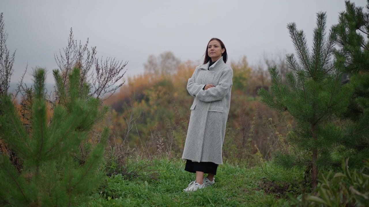 Ritratto a figura intera di donna in piedi da sola in un campo autunnale incolto, braccia incrociate e testa inclinata verso l'alto, che indossa un lungo cappotto chiaro, una gonna scura e stivali sotto un cielo nuvoloso e soffuso.
