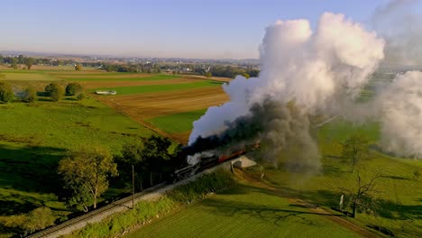 Vista-Aérea-De-Las-Tierras-De-Cultivo-Al-Amanecer-Con-Un-Motor-De-Vapor-Y-Un-Tren-De-Pasajeros-Acercándose-Con-Una-Cabeza-Llena-De-Vapor-Y-Humo