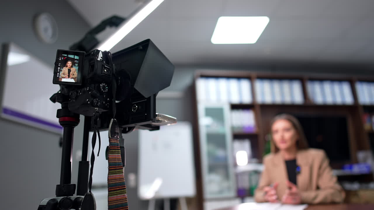 Display of camera on tripod shows a brunette woman in beige jacket. Low angle view. Blog creation in the office.