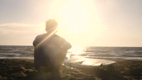 young man surfer in wetsuit sitting with surf board lying down on the sand at the beach listening to music dancing looking at the ocean at sunset