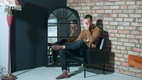 tired man sitting in a black chair near the mirror. man holds his head and rubs his face.