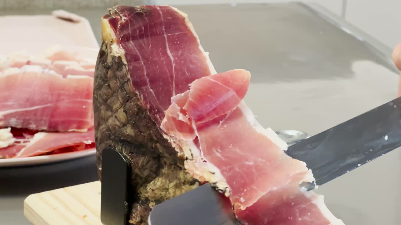 A piece of Jamón Ibérico (Spanish ham) being cut with a knife and a plate of Iberico cut meat in the background