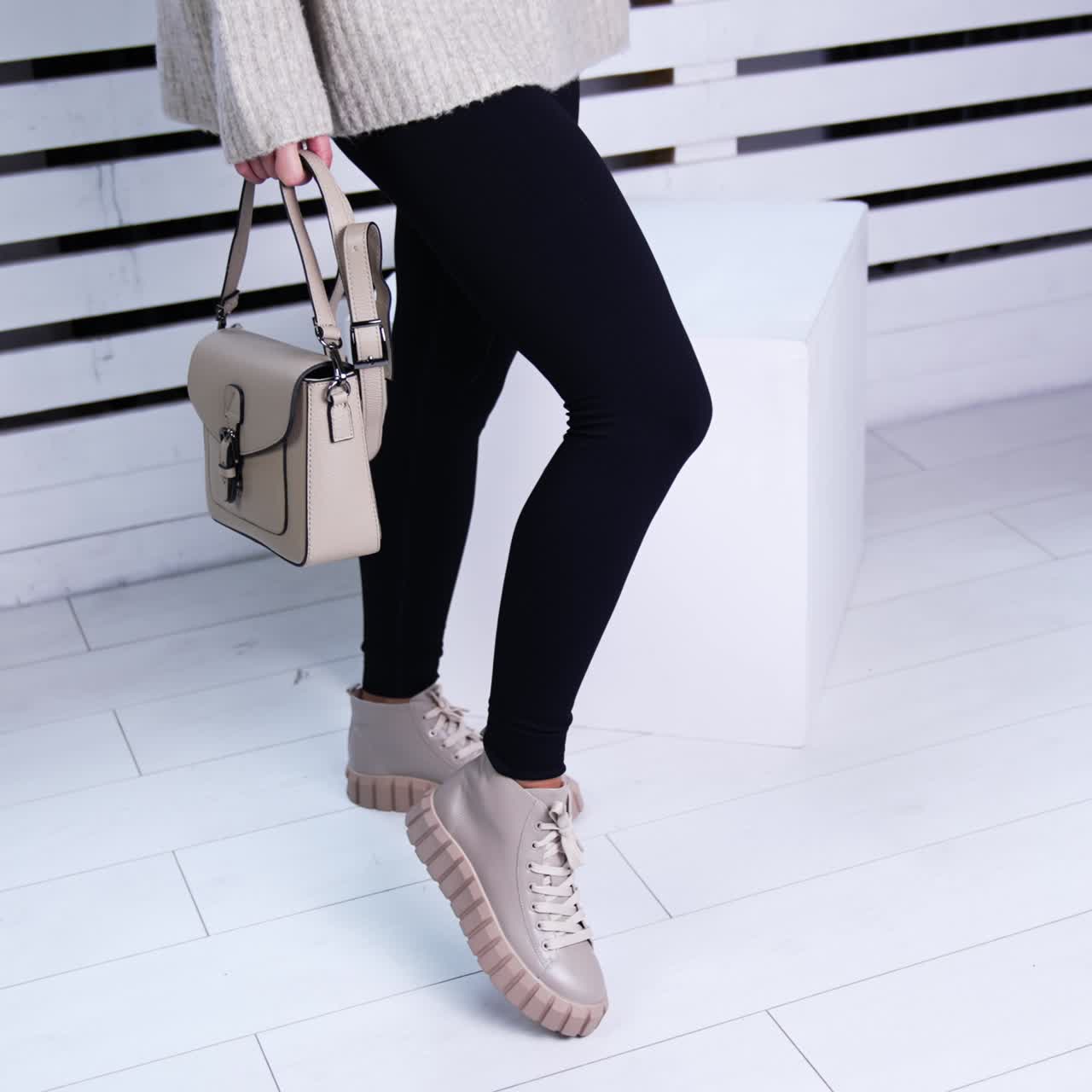 Woman in black tight pants, warm sweater and boots standing in the room. Lady with beige bag demonstrating sneakers. White backdrop