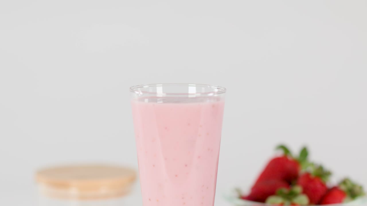 A hand places a strawberry slice atop a creamy pink milkshake under bright studio lighting